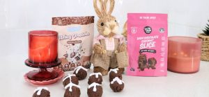 Front on photo of Whey Protein by Feel Good Protein (Chocolate Milk) sachet and a Rich Chocolate Coconut Slice Mix by Get Ya Yum On sachet on both sides of a Easter bunny statue centred behind 8 protein balls.