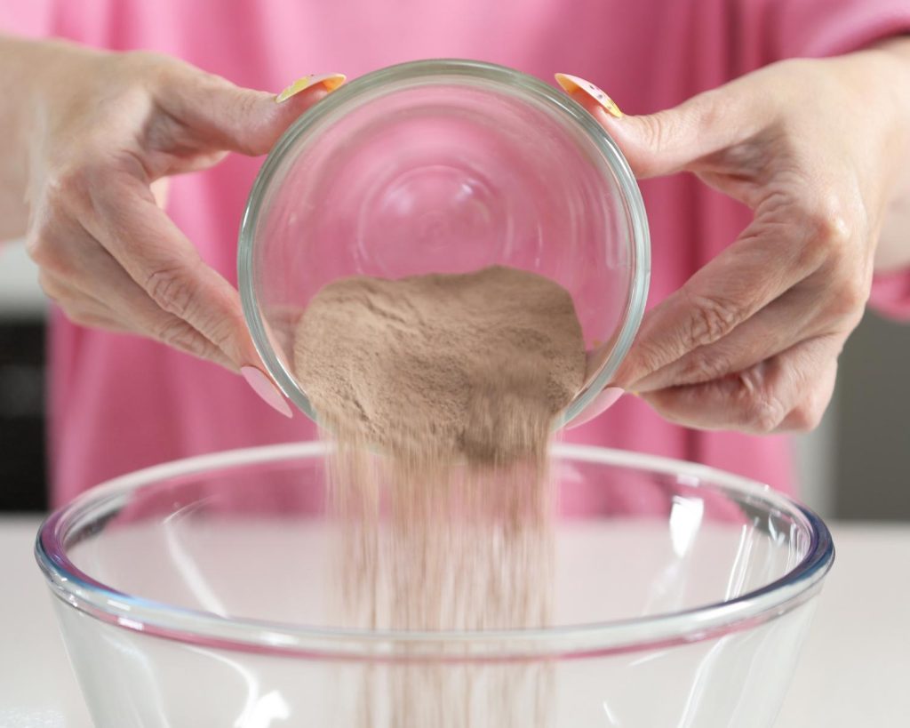 Close-up photo of two hands pouring a small bowl of brown powder into a larger clear glass mixing bowl.