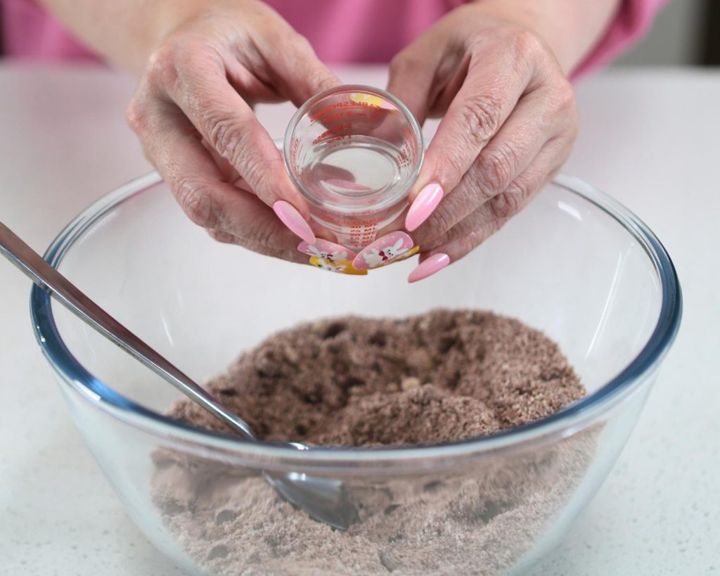 Close-up photo of two hands holding a small measuring glass of water over a large clear glass mixing bowl half filled with dry ingredients.