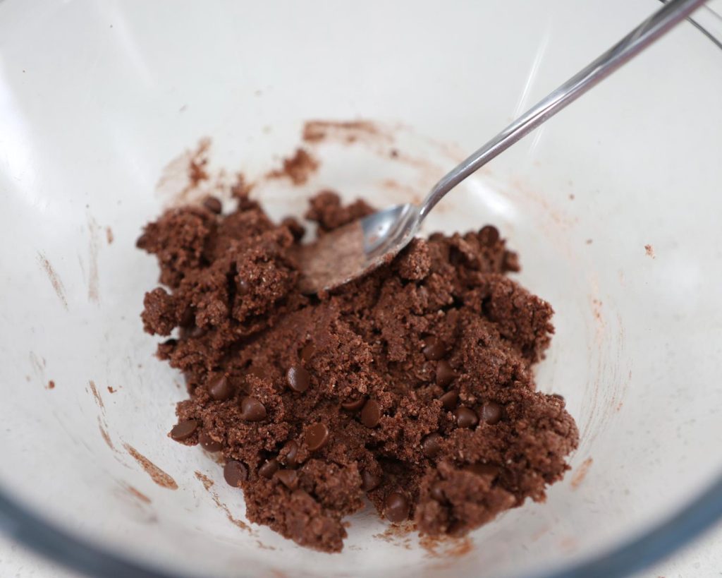 Close-up of protein ball mixture ingredients mixed in a clear glass mixing bowl with a metal spoon resting on the right side.
