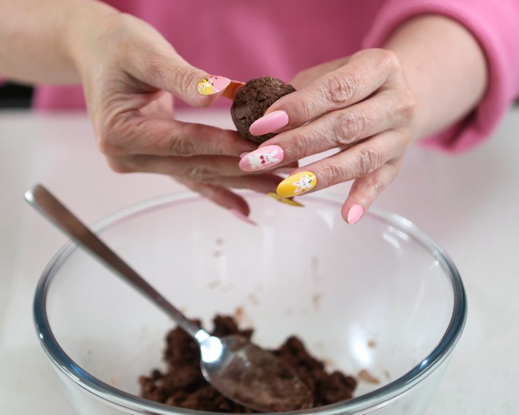 Close-up photo of two hands shaping a protein ball over a large clear glass mixing bowl half filled with protein ball mixture.