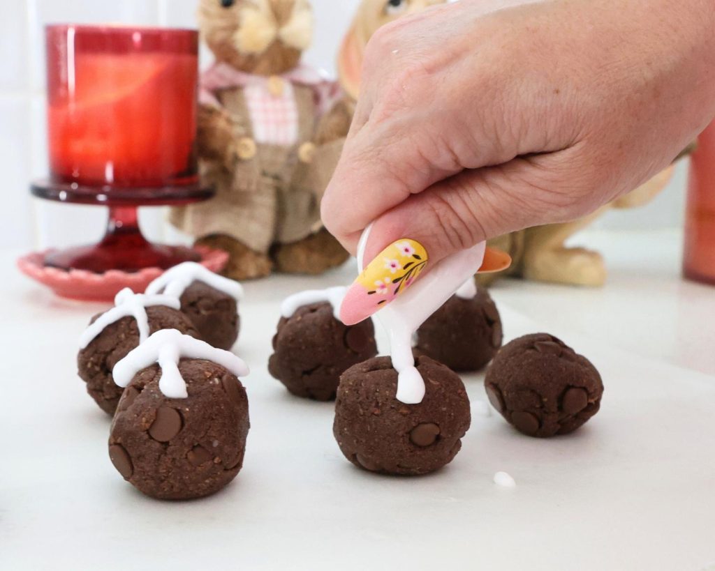 Close-up of a hand pipping icing into crosses on top of a protein ball, surrounded by 6 other protein balls in the background.