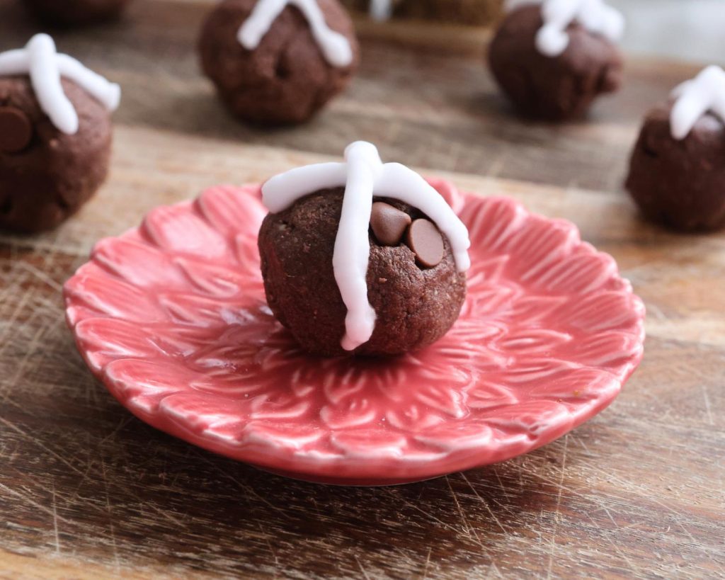 Close-up photo a hot cross bun protein ball placed on a small red plate with flower patterns, in the foreground of 6 hot cross bun protein balls resting on a wooden board.