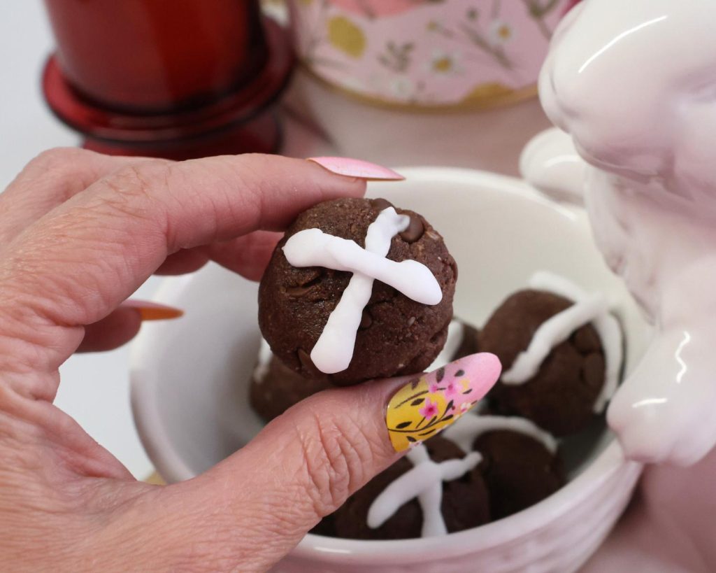 Close-up photo of a hand holding a hot cross bun protein ball p in the foreground of a small white bowl filled with hot cross bun protein balls.