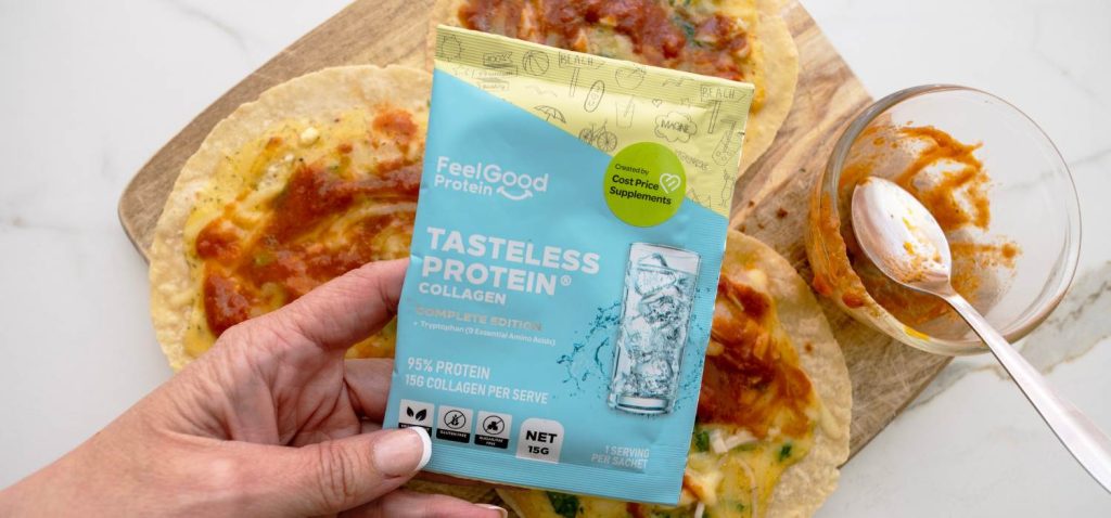 Top-down photo of a hand holding a Tasteless Protein Collage by Feel Good Protein sachet over the centre of a wooden board with three pizzas, a small glass bowl, and spoon on a white surface.