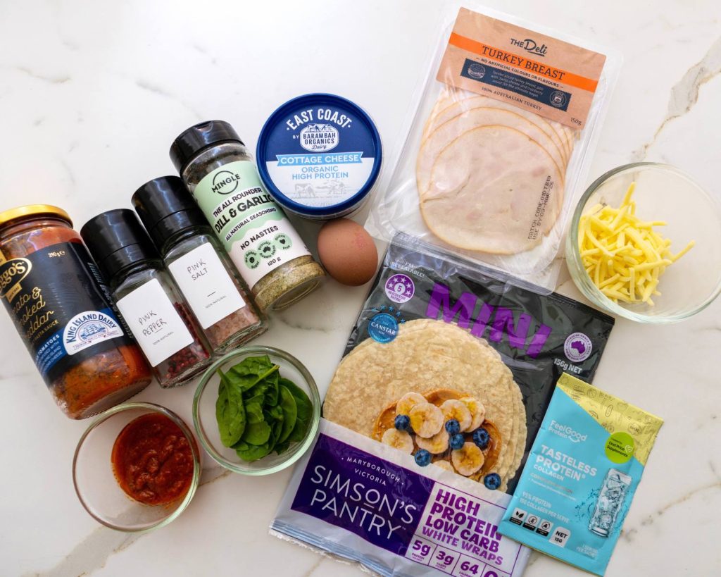 Top-down photo showing all the ingredients for high protein breakfast pizza wraps arranged side by side on a benchtop. 