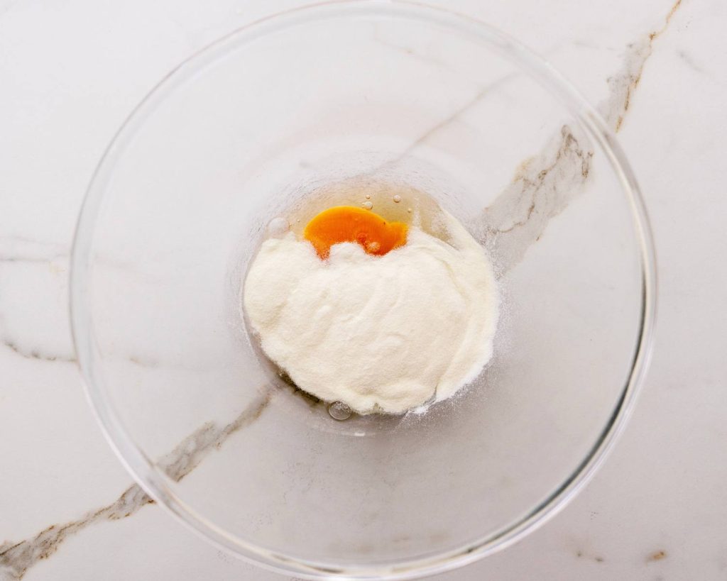 Overhead view of a clear glass mixing bowl half‑filled with protein powder and eggs in view, on top of a white benchtop. 