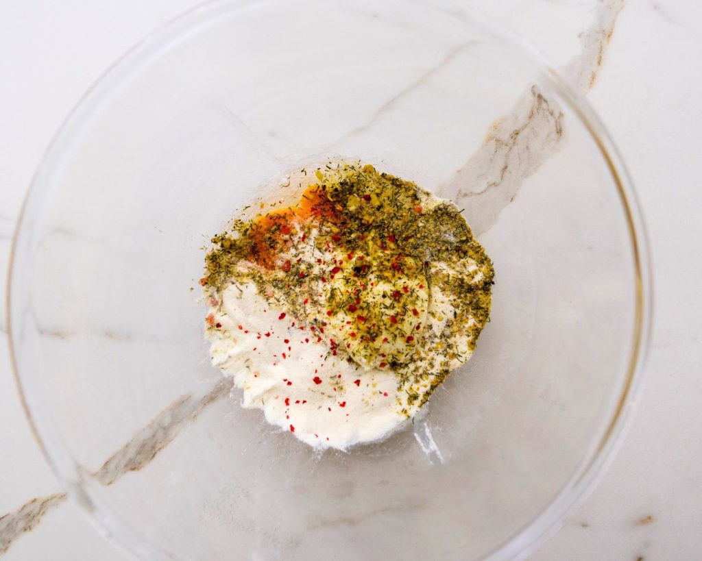 Overhead view of a clear glass mixing bowl half‑filled with protein powder, eggs, and spices in view, on top of a white benchtop. 