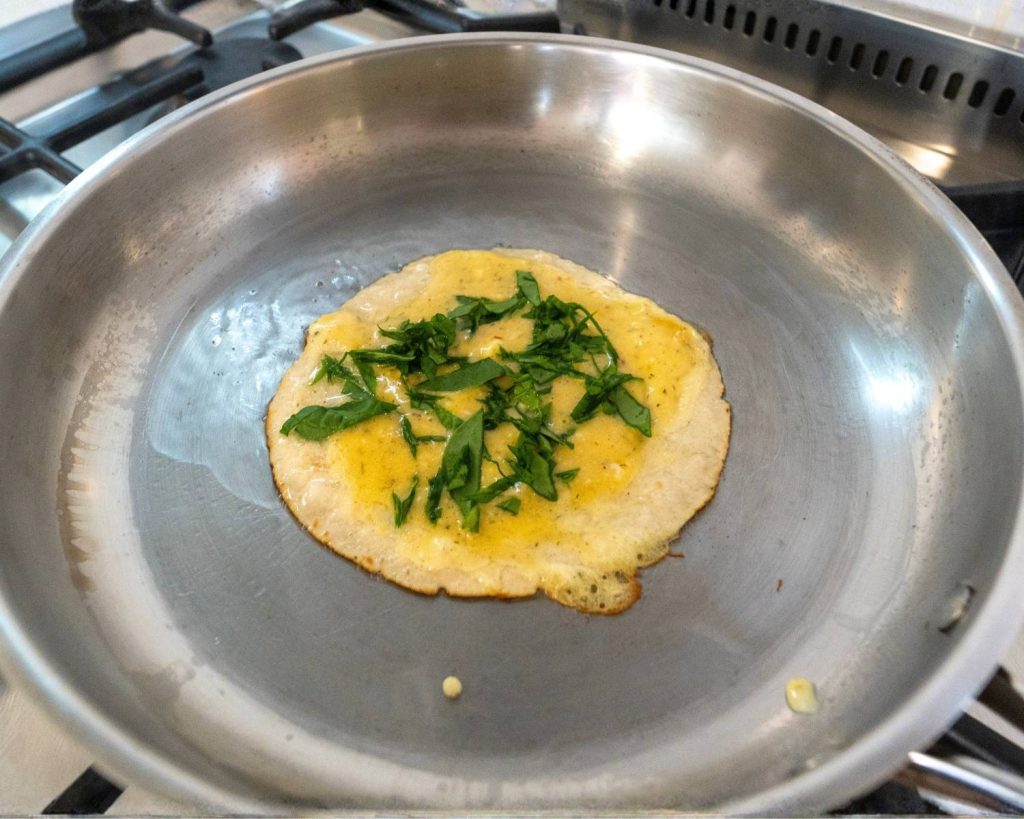 Point of view photo of a silver pan on a stovetop with breakfast pizza mid cooking, on top of a white benchtop. 