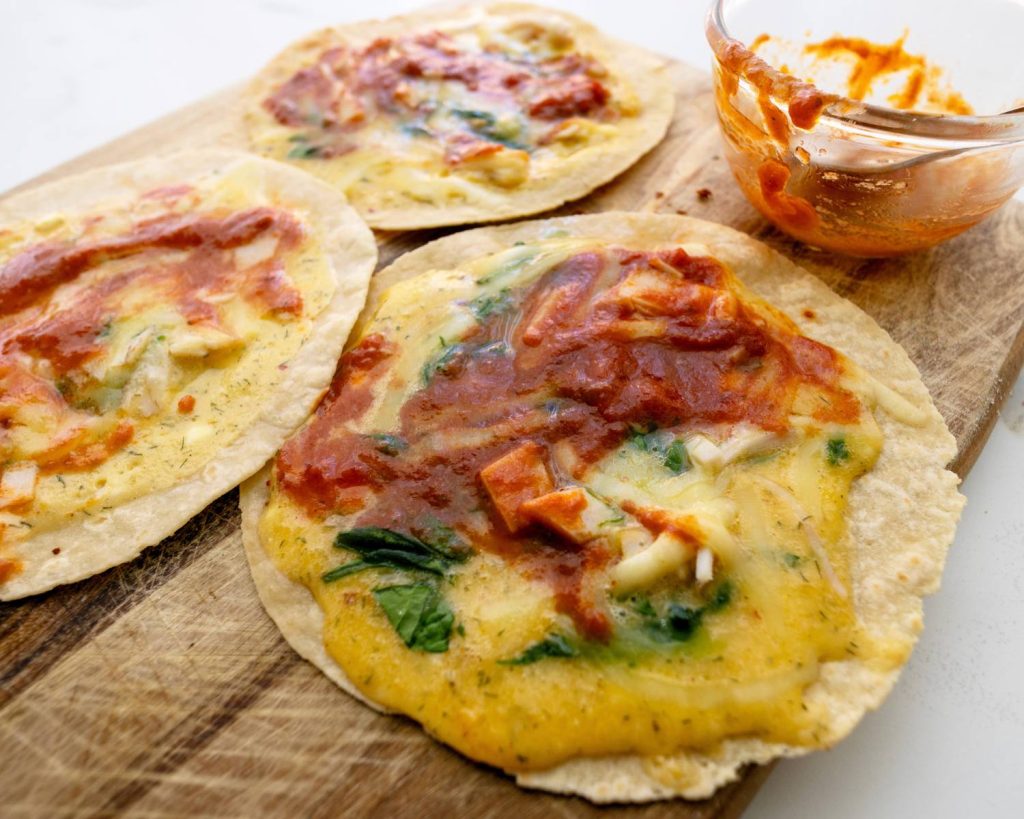 Point of view photo 3 breakfast pizza wraps, and a small glass bowl placed on across a wooden board in front of a blurred benchtop background. 