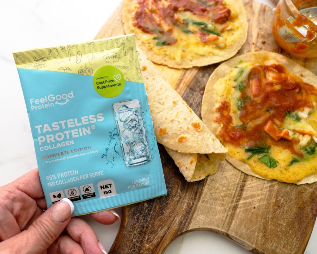 Point of view photo of hand holding a yellow, blue, and white Tasteless Protein Collagen by Feel Good Protein sample sachet in front of 3 breakfast pizza wraps and a small glass bowl, placed on across a wooden board in front of a blurred benchtop background. 