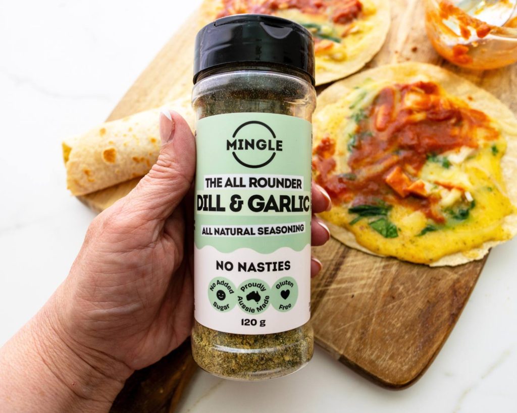 Point of view photo of hand holding a Mingle seasoning bottle in front of 3 breakfast pizza wraps and a small glass bowl, placed on across a wooden board in front of a blurred benchtop background. 