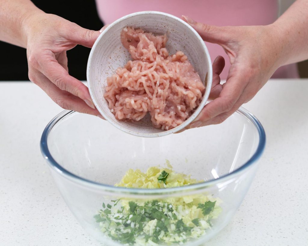 Close-up photo of two hands holding a small white bowl filled with chicken mince over a large clear glass mixing bowl half filled with vegetables.