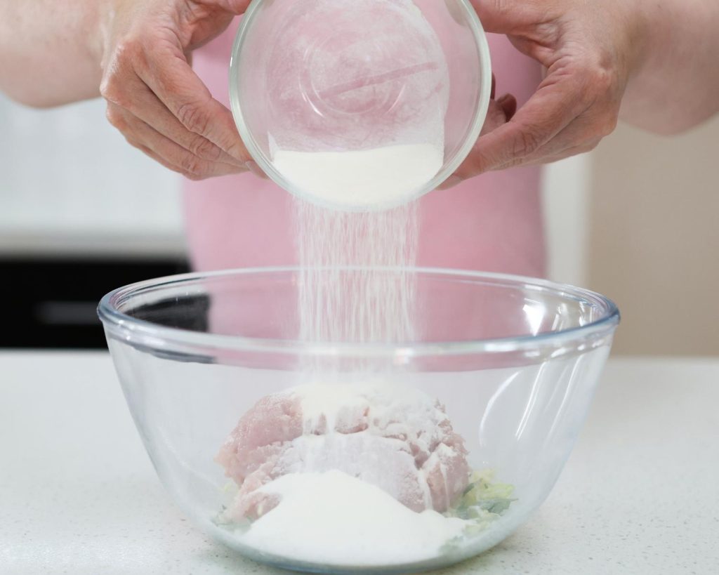 Close-up photo of two hands holding a small glass bowl filled with protein powder over a large clear glass mixing bowl half filled with chicken mince and vegetables.