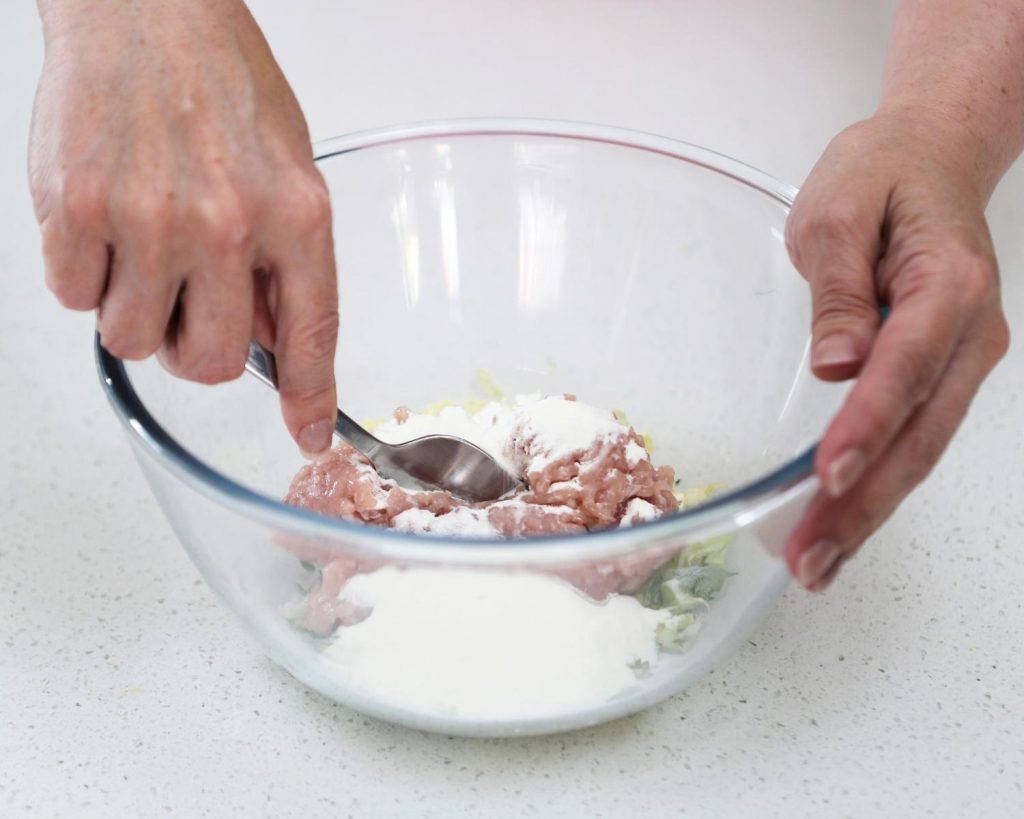Close-up photo of a hand using a metal spoon to mix parcel filling ingredients in a large clear glass mixing bowl.