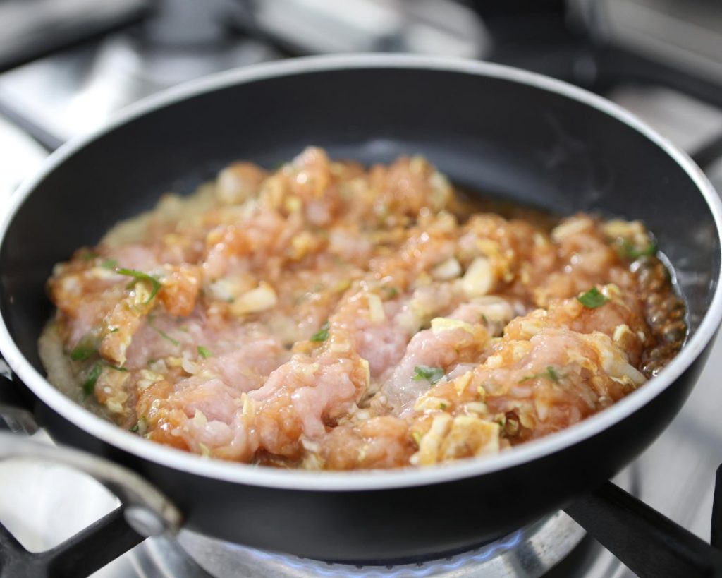 Point of view photo of a silver and black pan on a stovetop with chicken mince filling mixture cooking, on top of a benchtop.