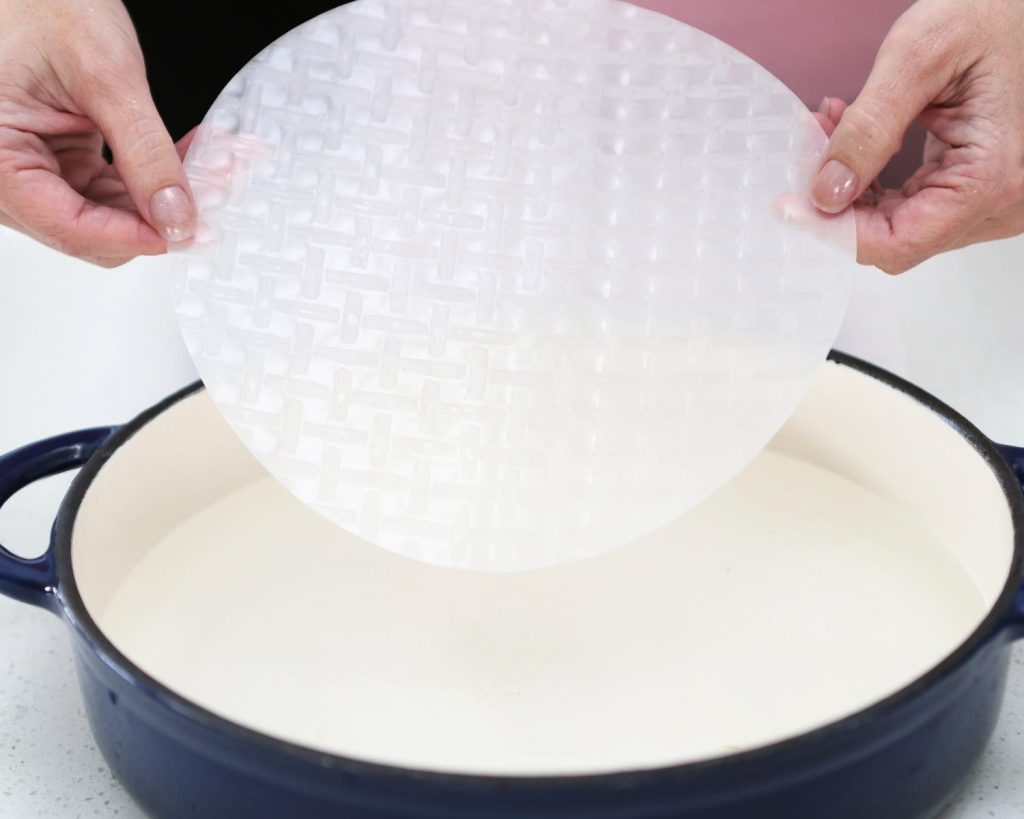 Point of view photo of two hands holding a circular rice paper sheet above a black pot filled with water.