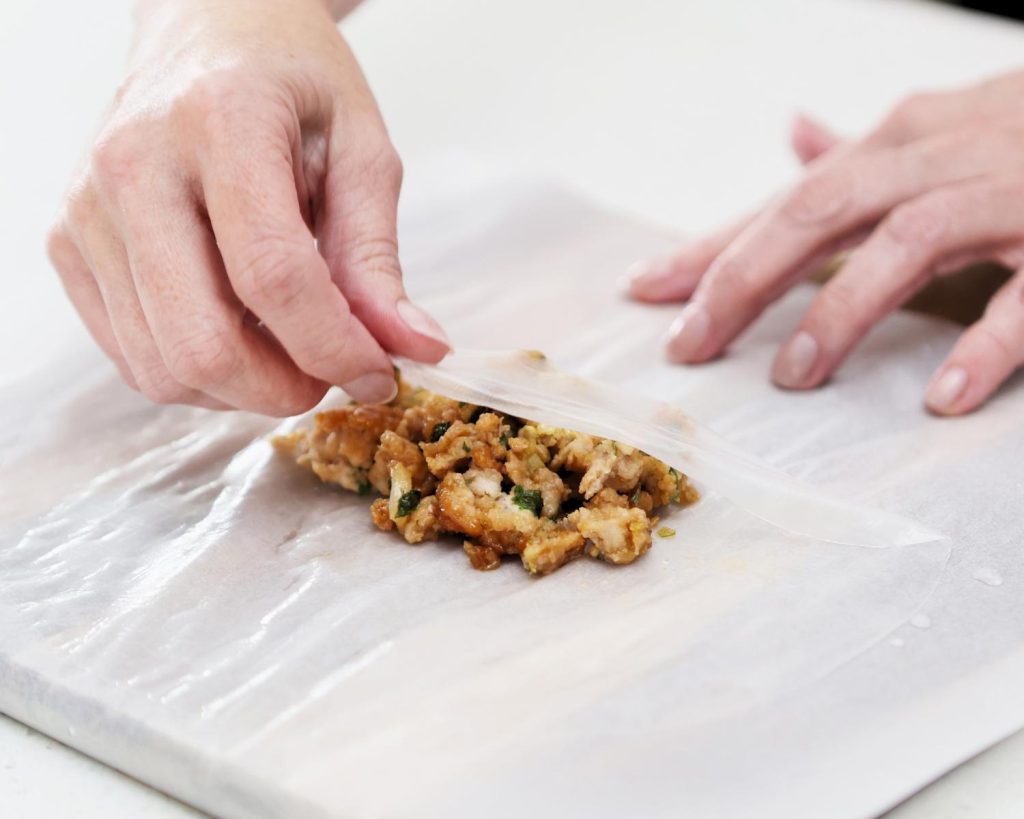 Point of view photo of two hands folding a circular rice paper sheet over a small pile of cooked chicken mince the centre of a board.