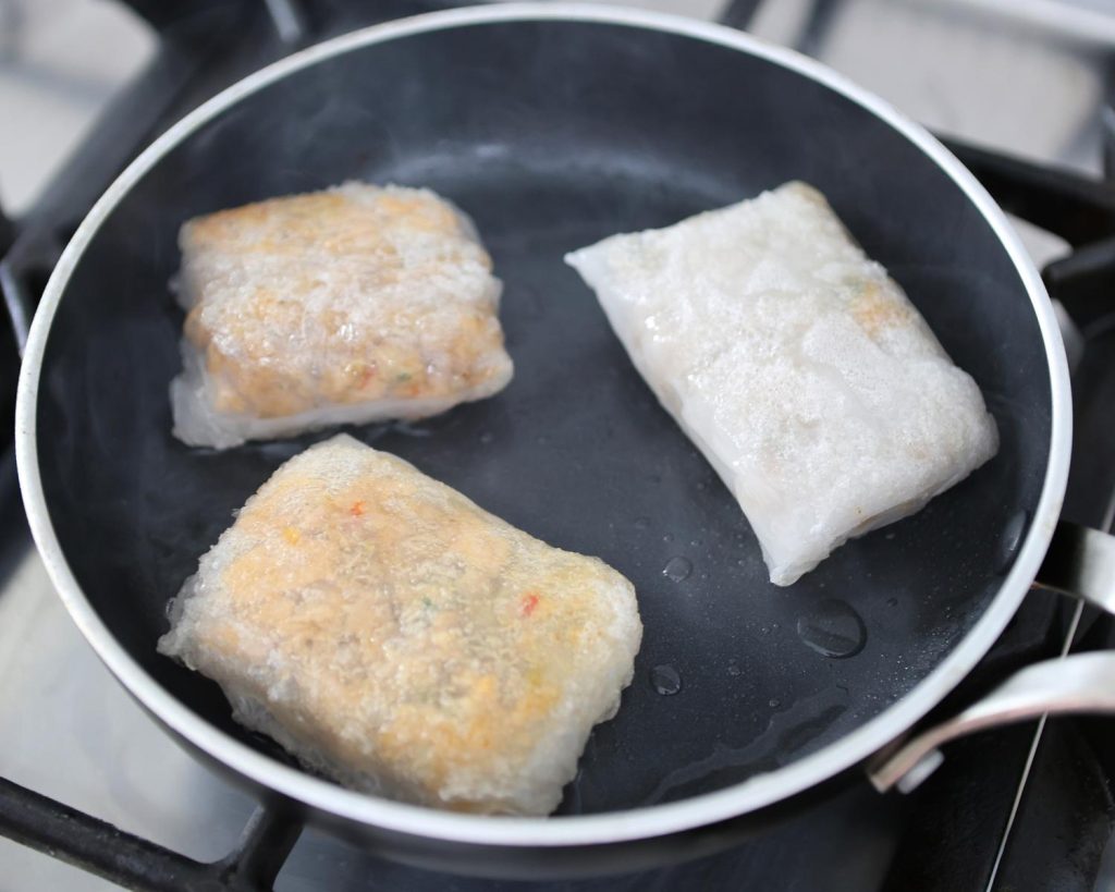 Point of view photo of a silver and black pan on a stovetop with 3 rice paper parcels cooking, on top of a benchtop.