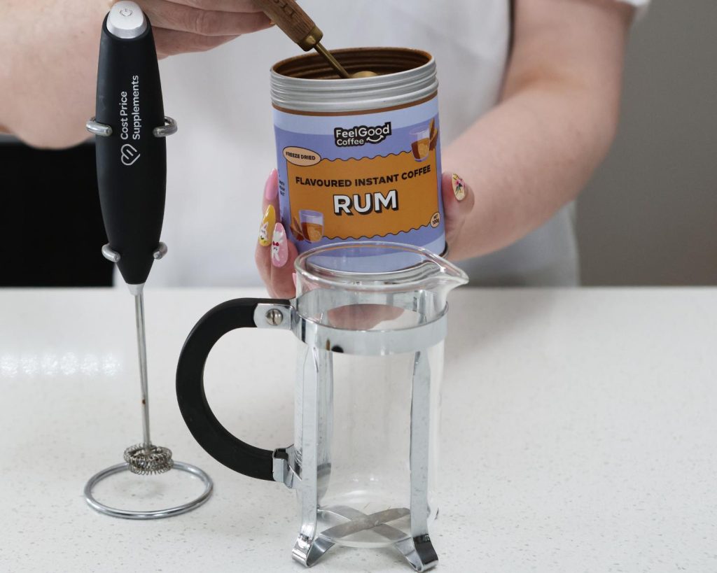 Front-facing photo of two hands scooping instant coffee out of a metal cannister with orange and purple branding reading ‘Flavoured Instant Coffee Rum’, positioned behind a black metal Cost Price Supplements electric whisk and a glass coffee press. 
