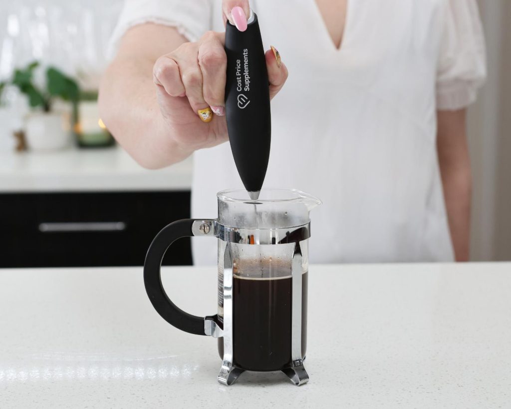 Front-facing photo of a hand holding a metal and black Cost Price Supplements electric whisk in a glass coffee press filled with coffee. 