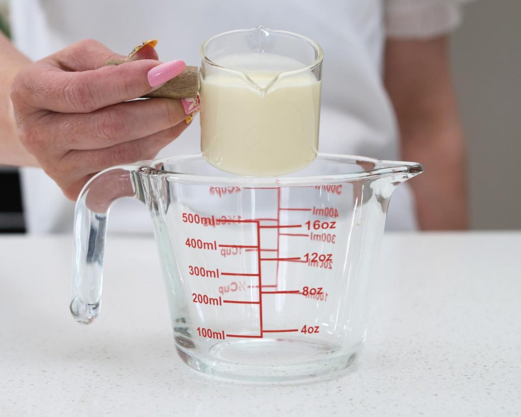 Front-facing photo of a hand pouring a small glass measuring cup of cream into an empty measuring jug. 
