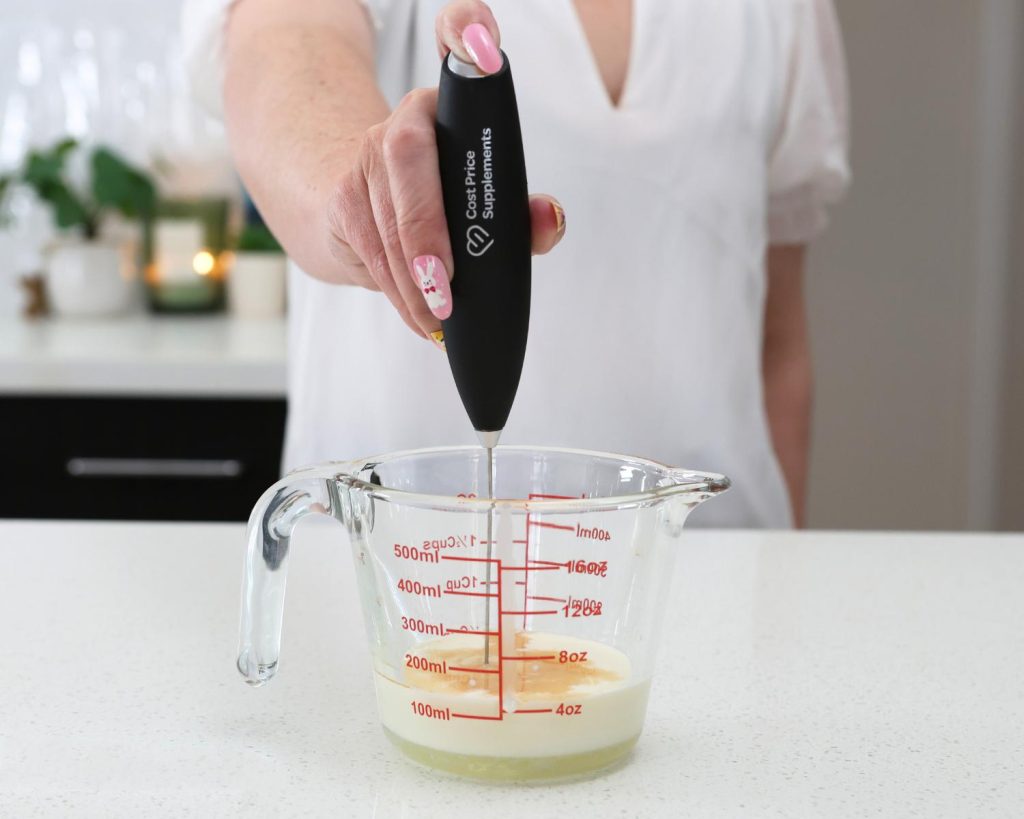 Front-facing photo of a hand holding a metal and black Cost Price Supplements electric whisk in a glass measuring jug filled with cream. 