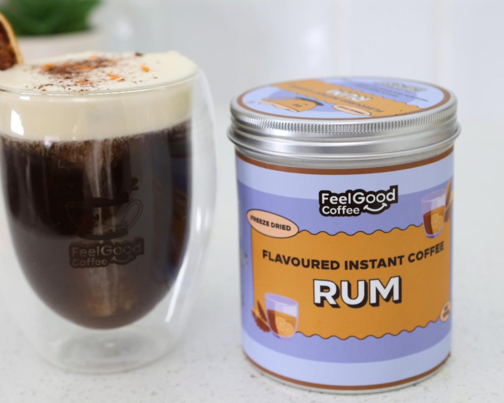 Front-facing photo of a Mont Blanc Proffee in a glass coffee cup on the left of a metal cannister with orange and purple branding reading ‘Flavoured Instant Coffee Rum’. 