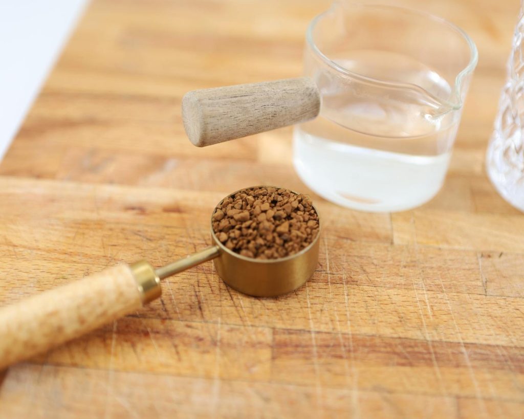 Top-down photo of a metal and wooden tablespoon filled with instant coffee, next to a glass measuring jug filled with water, resting on a wooden board.  