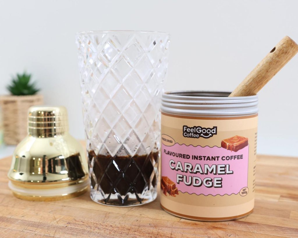 Front-facing photo of coffee in a glass cocktail shaker on the left of a metal cannister with pink and yellow branding reading ‘Flavoured Instant Coffee Caramel Fudge’. 