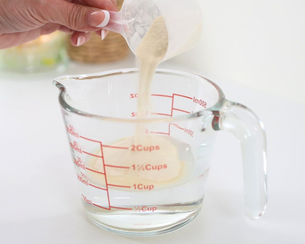 Front-facing photo of a hand pouring a scoop of protein powder into a glass measuring jug filled with water. 
