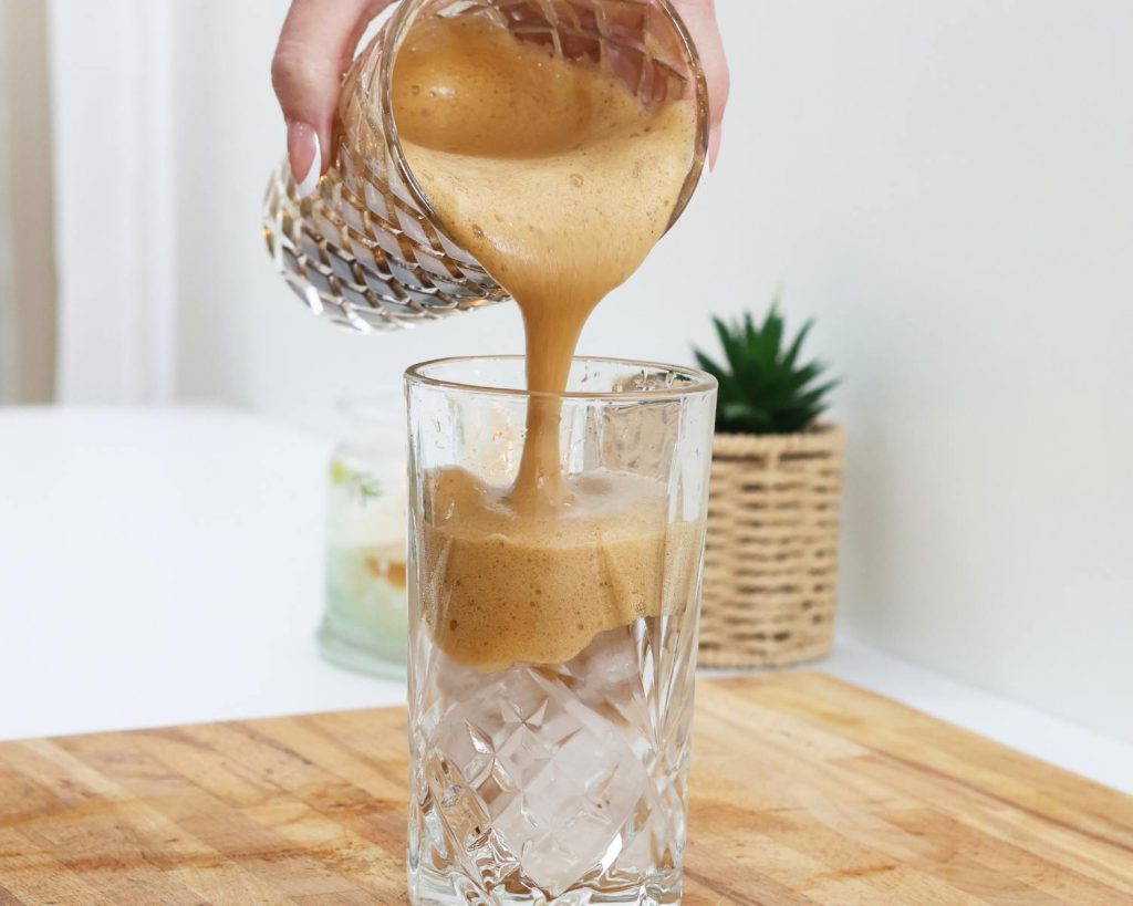 Front-facing photo of a hand pouring coffee into a glass cup filled with ice, resting on a wooden board. 