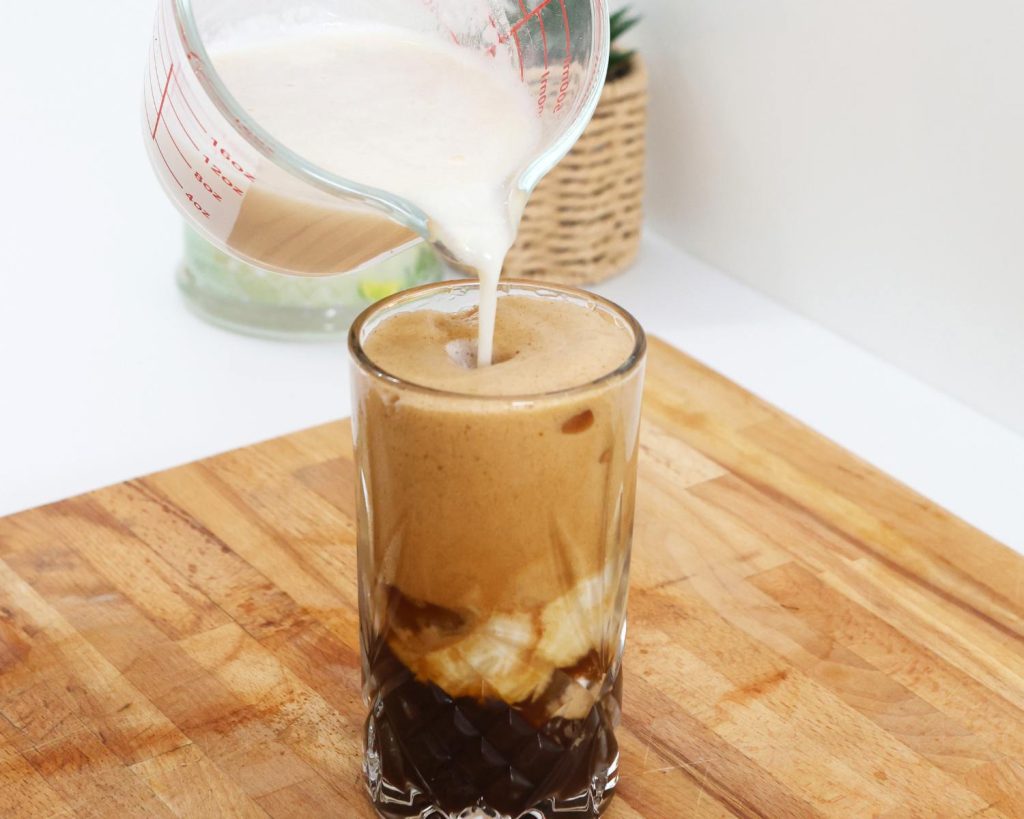 Front-facing photo of a whey mixture into a glass cup half filled with coffee and ice, resting on a wooden board. 