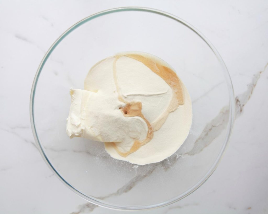 Overhead view of a clear glass mixing bowl half‑filled with protein powder and cream cheese in view, on top of a white benchtop. 