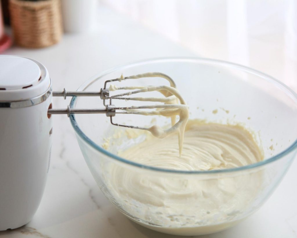 Photo of a clear glass mixing bowl half‑filled with No Bake High Protein Cheesecake Cups, with a hand mixer resting upright on the rim. 
