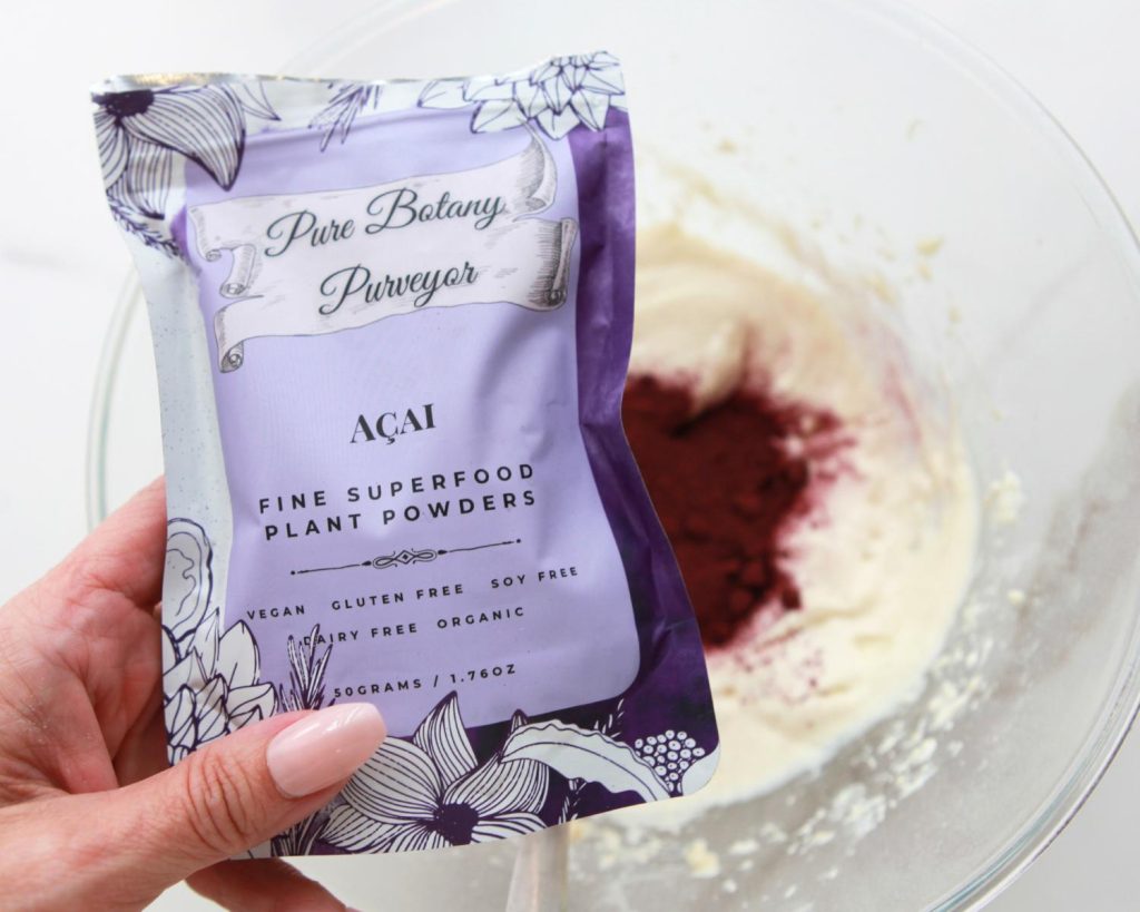 Overhead shot of a hand holding a purple branded Acai Fine Superfoods Powder by Pure Botany Purveyor bag in front of a blurred bowl of No Bake High Protein Cheesecake Cups mix. 