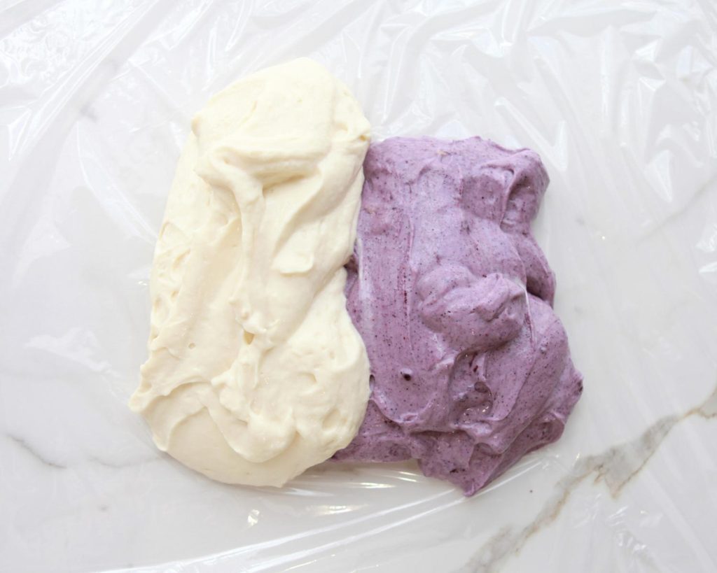 Overhead shot of purple and white No Bake High Protein Cheesecake Cups mixture spread out side-by-side on a benchtop covered with clingwrap. 