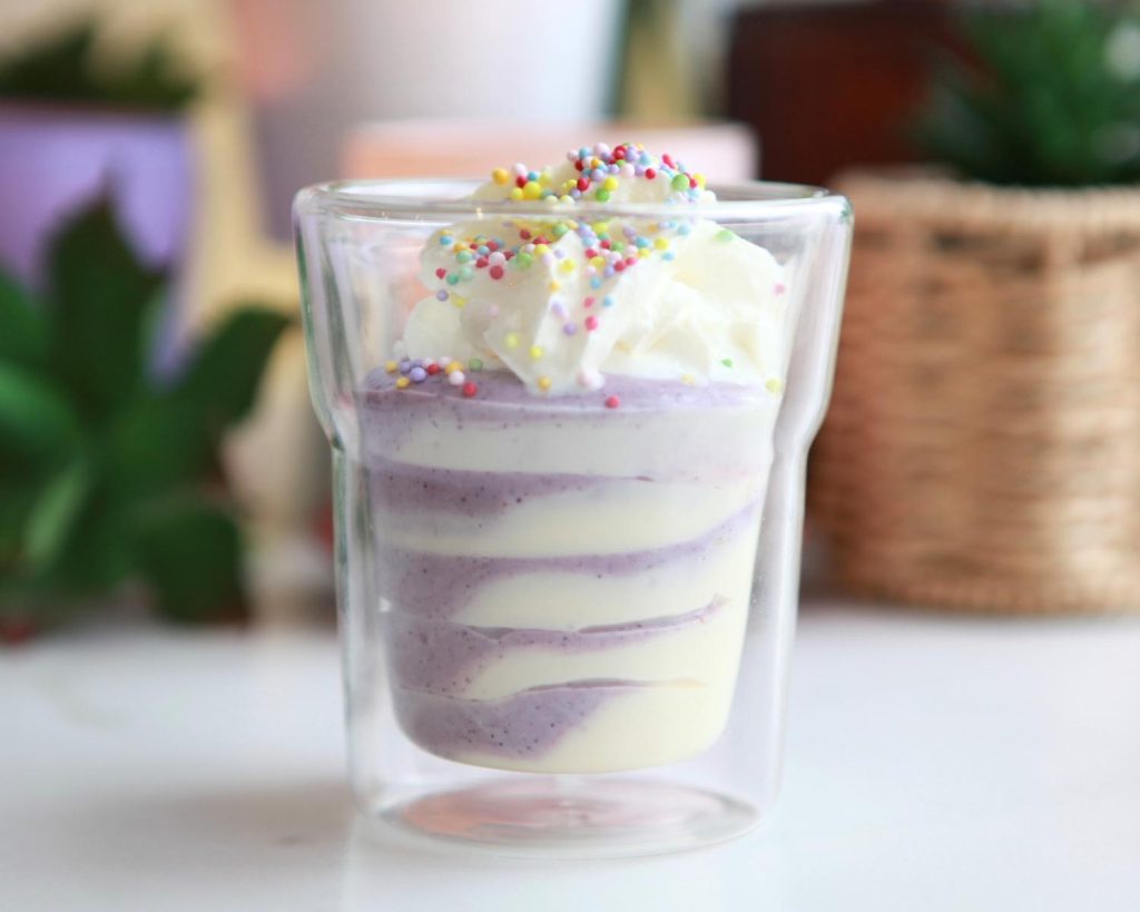 Close up photo of a small glass filled with No Bake High Protein Cheesecake Cup mixture, topped with whipped cream and rainbow sprinkles. 