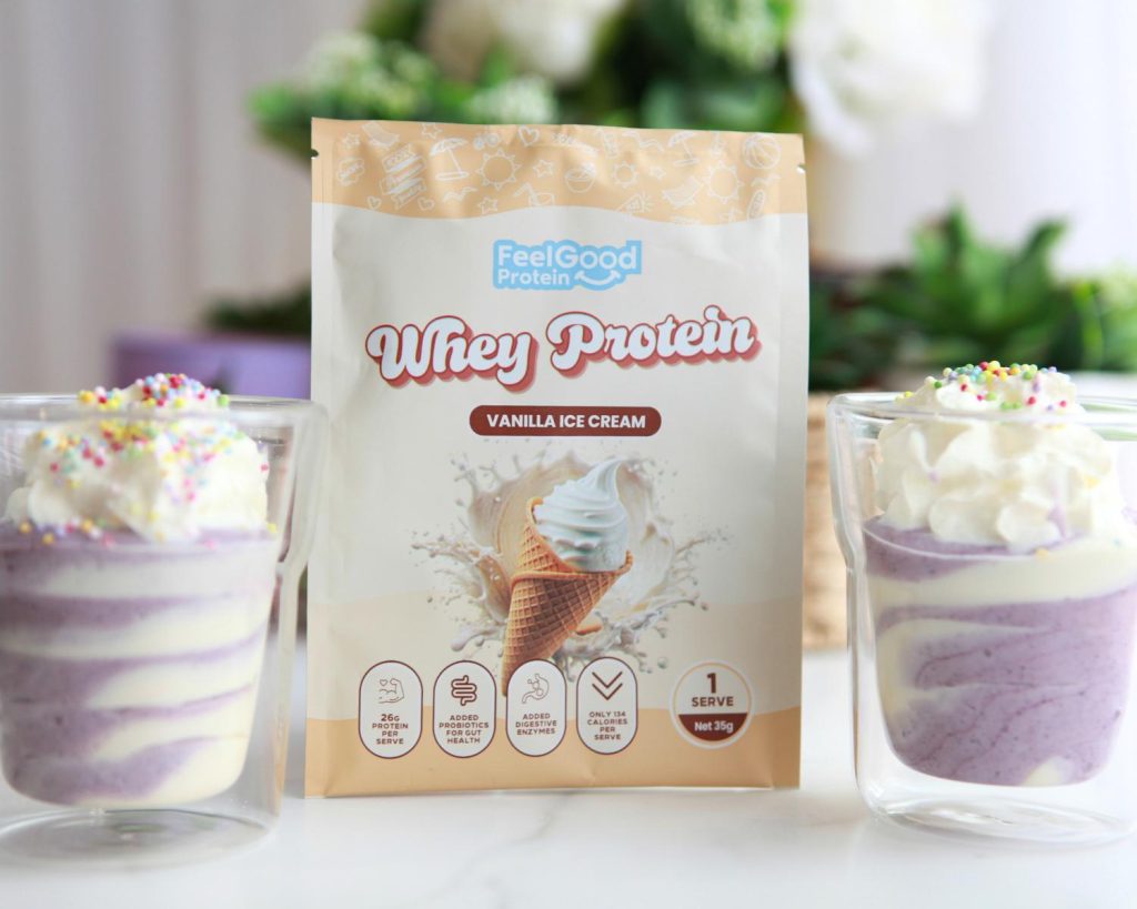 Front-facing photo of two No Bake High Protein Cheesecake Cups with a yellow and white sachet of Whey Protein by Feel Good Protein (Vanilla Ice Cream) in the centre, with a softly blurred modern kitchen in the background. 