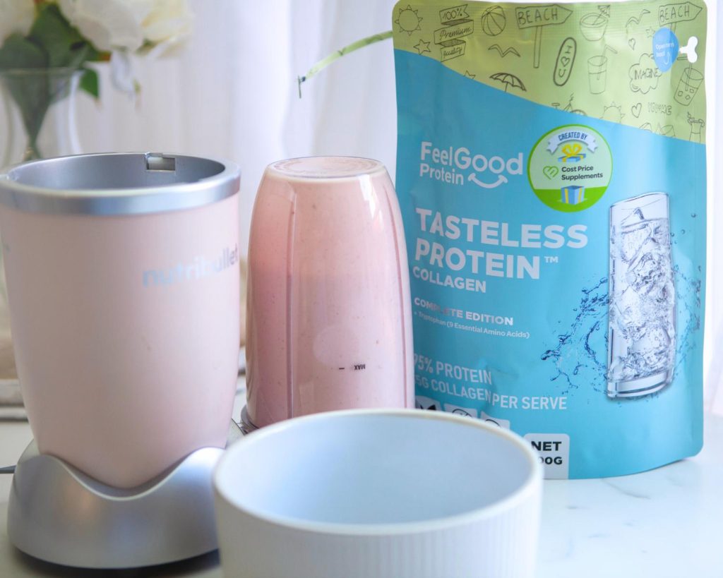 Front-facing photo of a pink blender half filled with Raspberry Swirl Protein Ice Cream mixture to the left of a full-sized Tasteless Protein Collage by Feel Good Protein bag, behind a large white bowl with a softly blurred modern kitchen in the background. 
