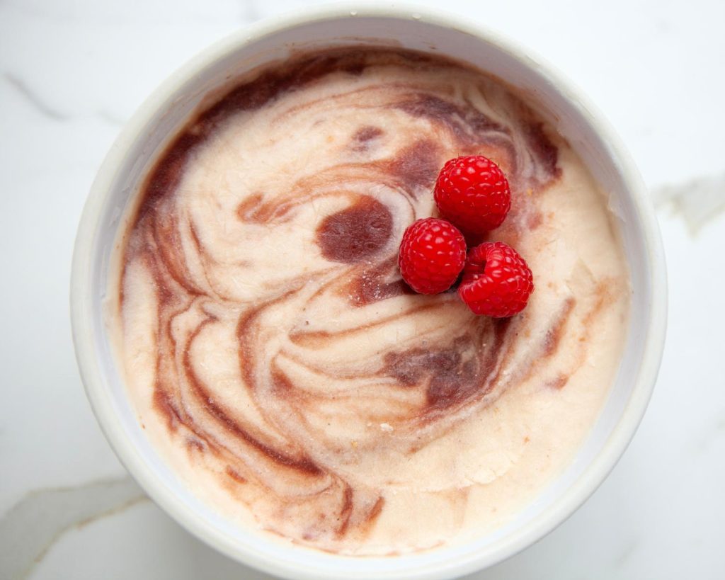 Top-down photo of a large white bowl filled with Raspberry Swirl Protein Ice Cream with three raspberries on top. 
