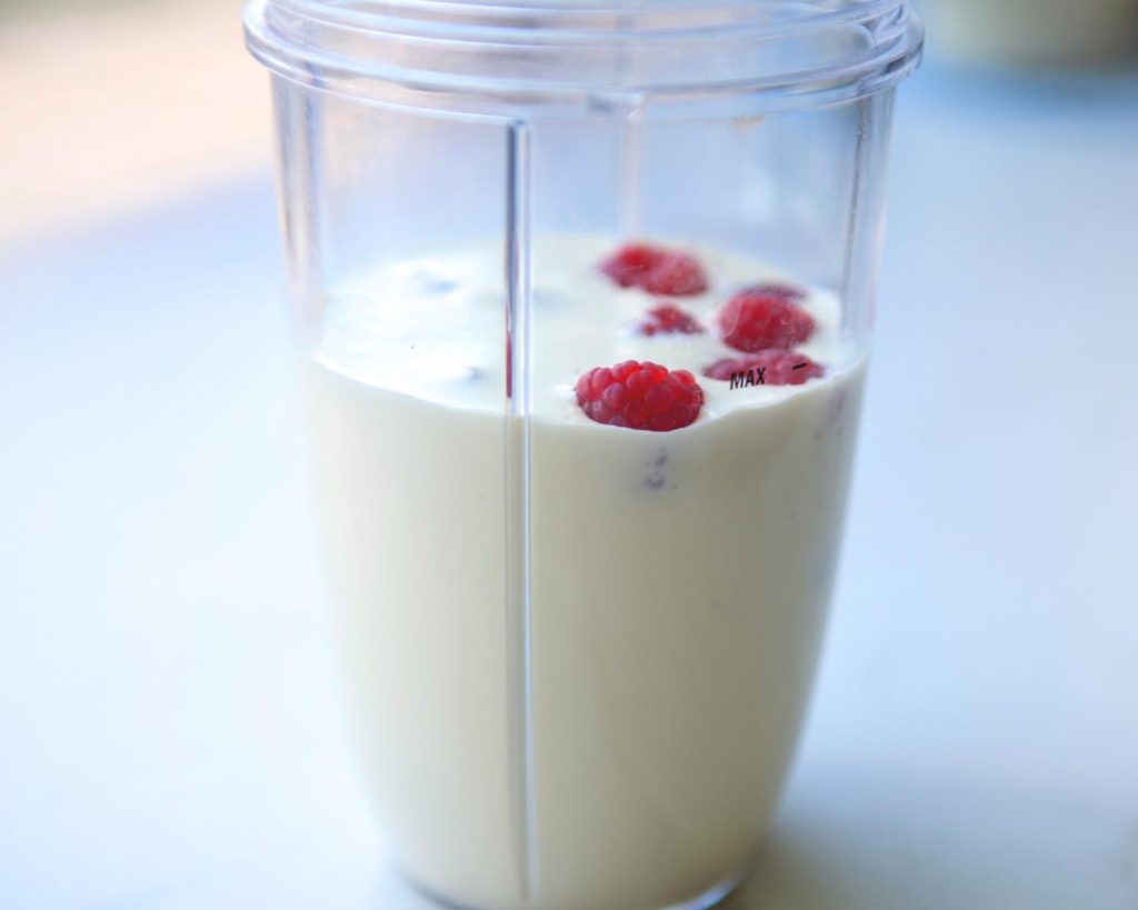 Front-facing photo of a clear blender half filled with raspberries and the liquid ingredients for Raspberry Swirl Protein Ice Cream with a softly blurred modern kitchen in the background. 