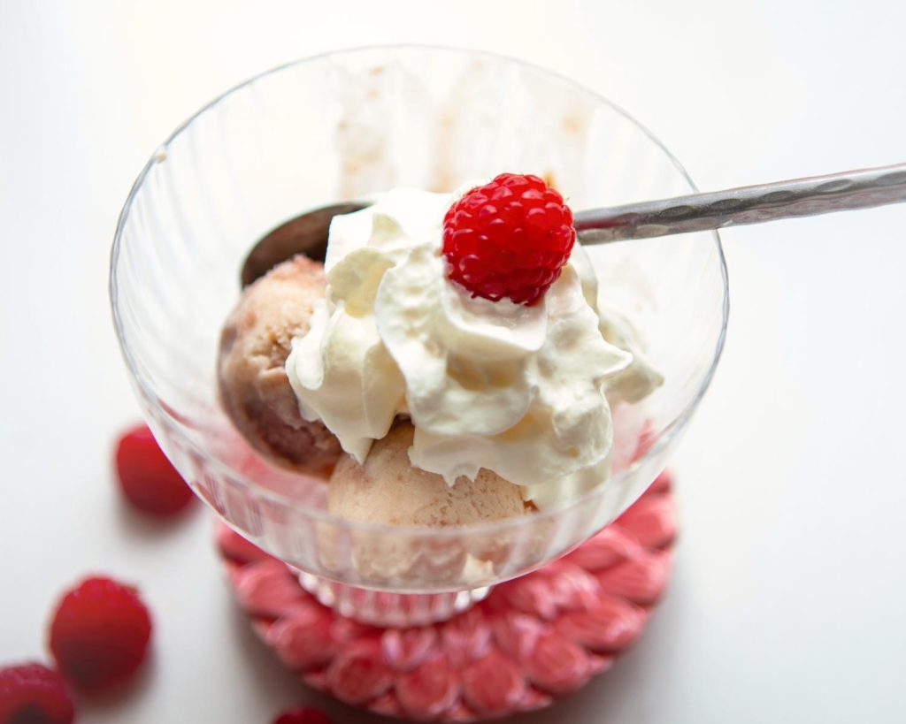 Top-down photo of a small glass bowl of Raspberry Swirl Protein Ice Cream, topped with whipped cream and raspberries. 