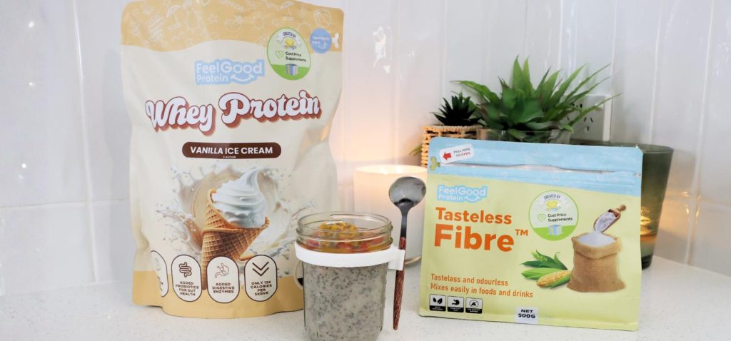 Front-facing photo of a glass container of High Protein Vanilla Ice Cream Chia Pudding in the centre of a blue and yellow sachet of Tasteless Fibre by Feel Good Protein and a yellow bag of Whey Protein by Feel Good Protein (Vanilla Ice Cream), with a softly blurred modern kitchen in the background.