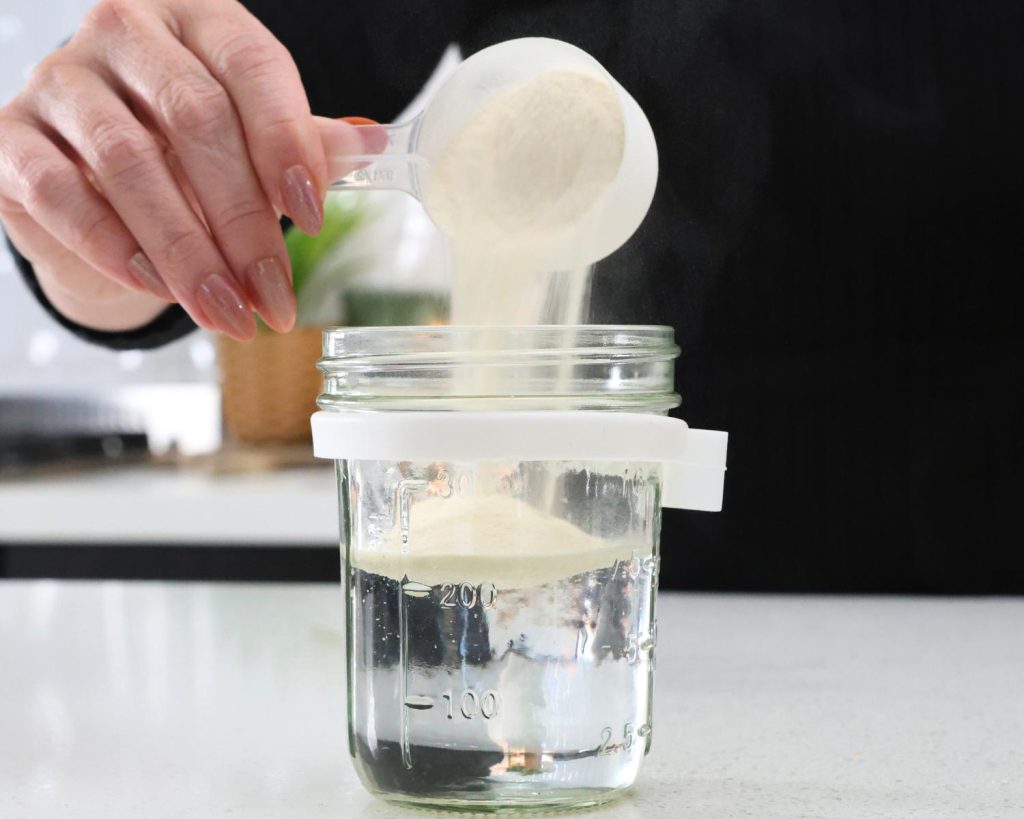 Front-facing photo of a hand pouring a scoop of Whey into a glass container filled with water. 