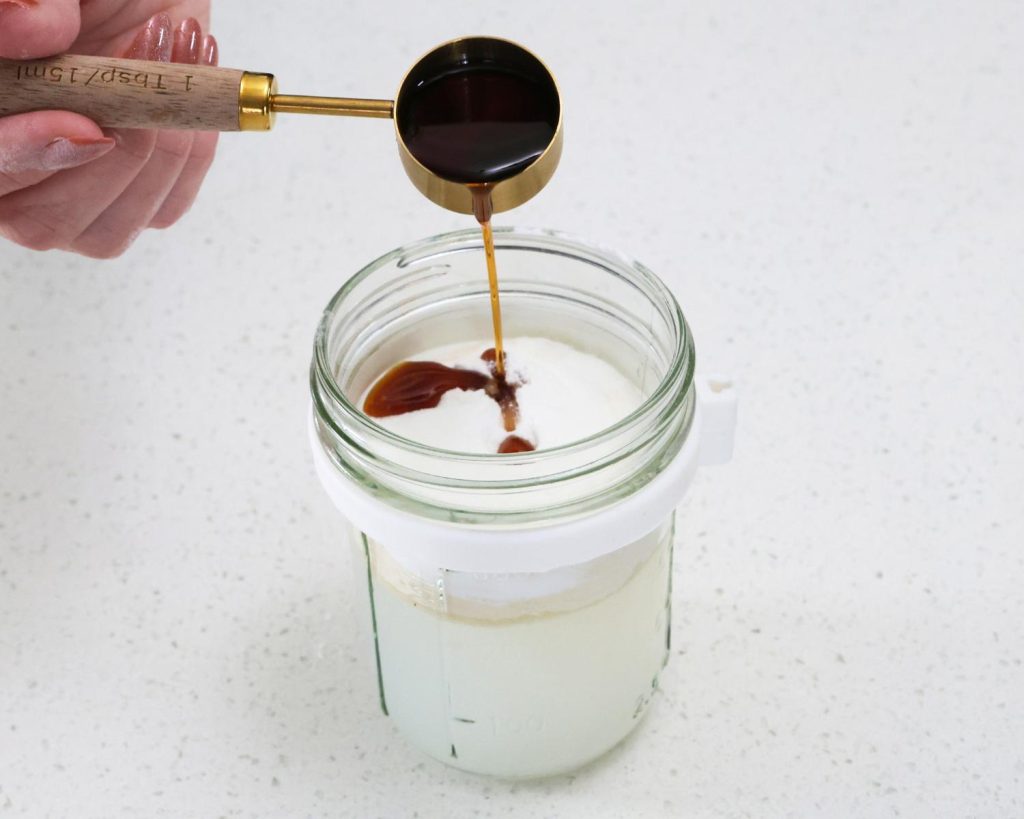 Front-facing photo of a hand pouring a tablespoon of vanilla extra into a glass container filled with water and whey. 