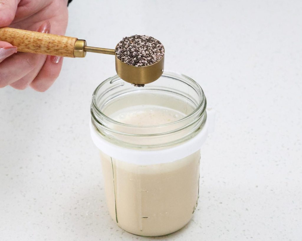 Front-facing photo of a hand pouring a tablespoon of chia seeds into a glass container filled whey mixture. 