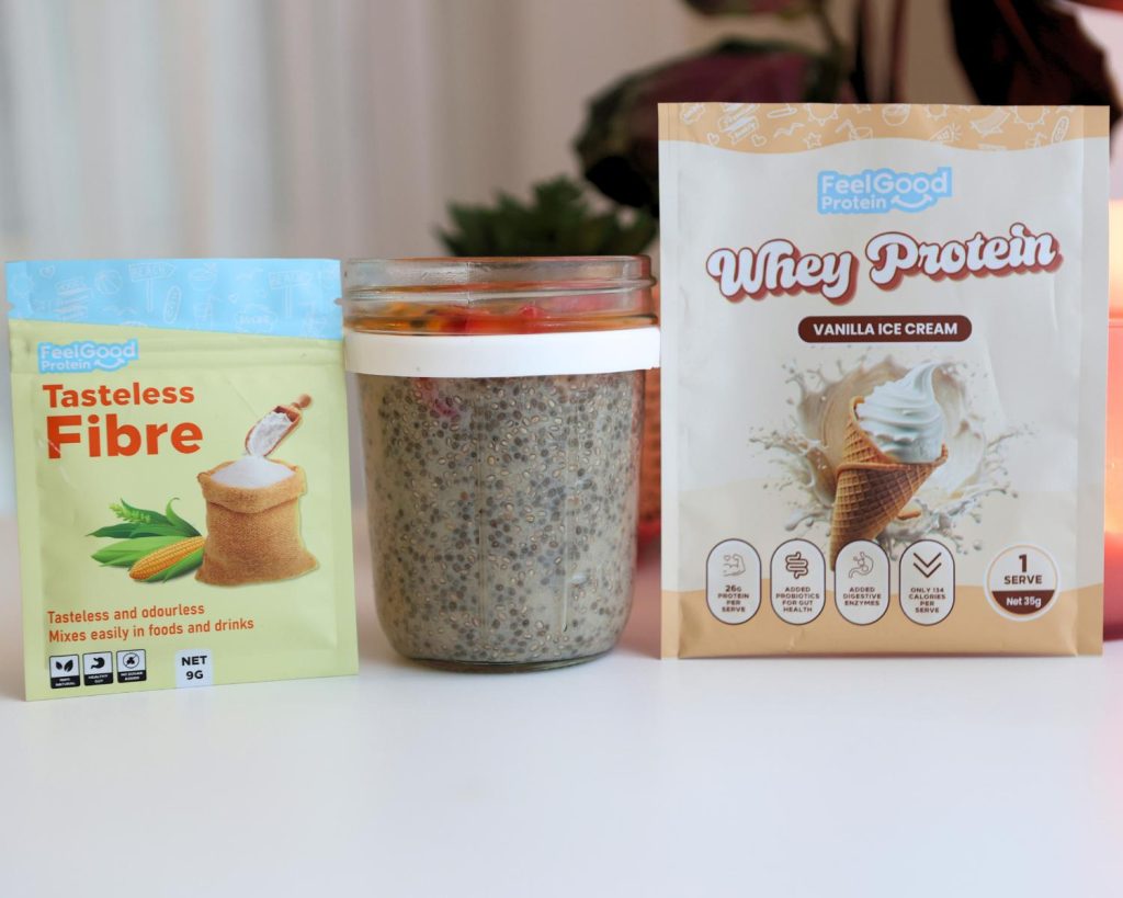 Front-facing photo of a glass container of High Protein Vanilla Ice Cream Chia Pudding in the centre of a blue and yellow sachet of Tasteless Fibre by Feel Good Protein and a yellow bag of Whey Protein by Feel Good Protein (Vanilla Ice Cream), with a softly blurred modern kitchen in the background. 