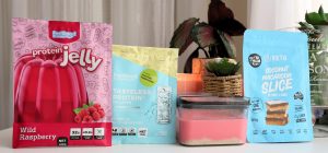 Front-facing photo of a container of High Protein Raspberry Cheesecake Slice in the centre of a blue and yellow sachet of Tasteless Protein Collagen by Feel Good Protein, a pink bag of Protein Jelly by Feel Good Protein (Wilf Raspberry), and a sachet of Coconut Macaroon Slice by Get Ya Yum On with a softly blurred modern kitchen in the background.