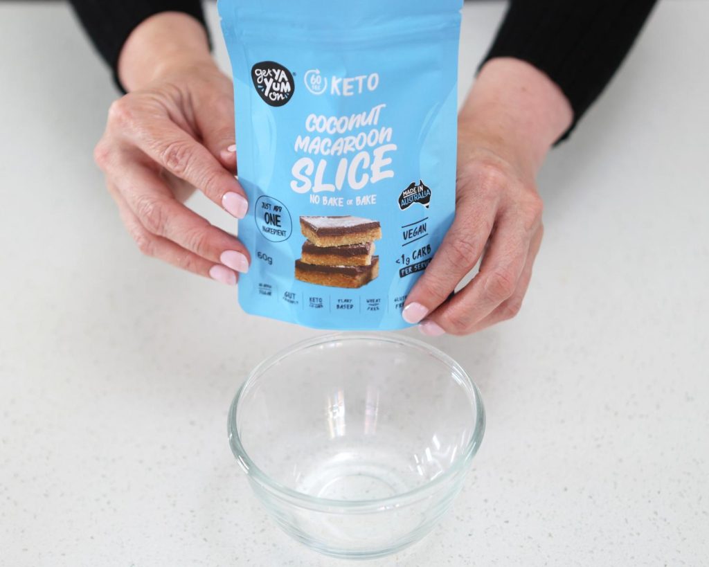 Front-facing photo of two hands holding a packet of Coconut Macaroon Slice by Get Ya Yum On over a glass mixing bowl.